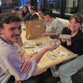 Group of people sitting around a table in a casual restaurant setting holding their pottery pieces. 
