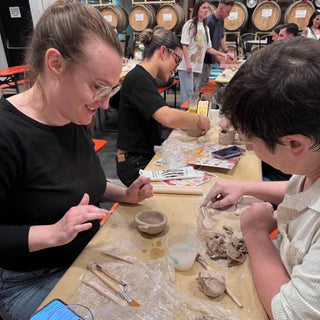 Three people working with clay at a pottery workshop with more people standing in the backgroung.