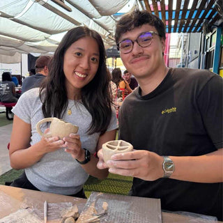 Two people holding ceramic pieces outdoors under a canopy.