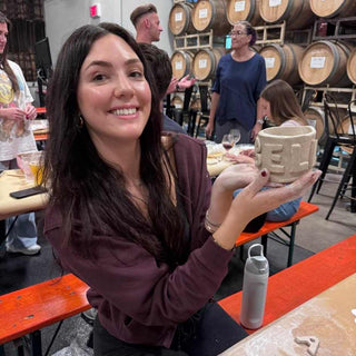 Woman holding a ceramic mug in a social setting with people and barrels in the background.