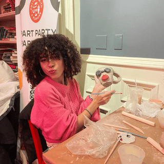 Woman in pink sweater holding a ceramic mug with a red handle at a pottery party event.