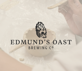 A hand holds a wooden spoon above a ceramic mug on a white surface, with the Edmund's Oast Brewing Co logo centered over the scene.