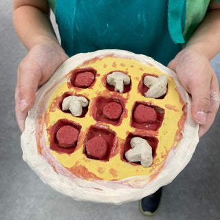 Model of a pizza with red and white toppings held by a person wearing a green apron.