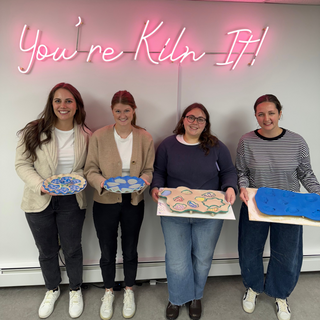 Four women holding ceramic oyster plates in front of a 'You're Kiln It!' neon sign.
