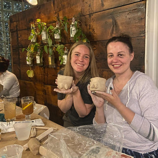 Two women holding ceramic cups in a pottery cafe with wooden walls and various pottery items.