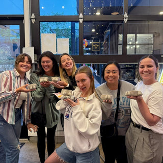 Group of six women posing together in a modern indoor setting holding clay pots they made