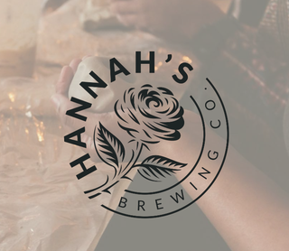 A circular black logo reading 'HANNAH'S BREWING CO' with a rose illustration over a softly blurred background of hands holding a mug.