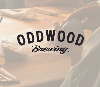 A warm-toned, slightly hazy photo showing a person holding a white mug at a wooden table, with the large text 'ODDWOOD Brewing' overlaid across the center.