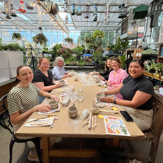 Group of people sitting around a table in a greenhouse setting, engaged in an sculpting pottery.