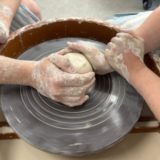 Close-up of a pottery wheel in use, with multiple hands shaping a lump of clay against the spinning wheel.