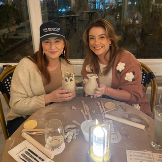Two women sitting at a table with art supplies and a candle, in a casual indoor setting.