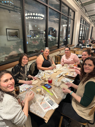 A group of five women sitting around a pottery table in a restaurant or workshop, holding and shaping clay pieces. The table is covered with pottery tools, water, and clay materials.