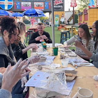 Group of people working on pottery at an outdoor event with various booths and umbrellas in the background.