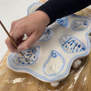 Person painting a ceramic dish with blue designs on a white surface.