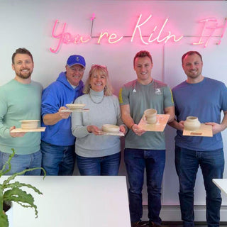 Group of people holding ceramic items in front of a neon sign with 'You're Kiln In' text.