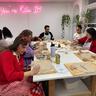 Group of people in a pottery studio with 'You're Kiln It!' neon sign.