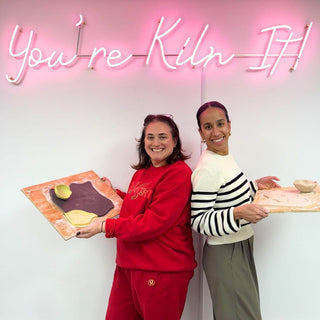 Two people holding trays with clay sculptures in front of a neon sign that reads 'You're Kiln It!'.