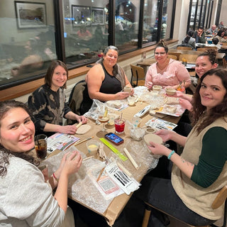Group of women sitting around a table in a casual setting, possibly a restaurant or cafe, scutping pottery.