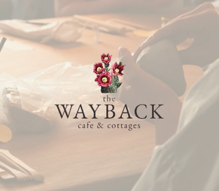 A warm-toned cafe interior scene with a person holding a white mug, overlaid with the text and floral logo 'the WAYBACK cafe & cottages'.