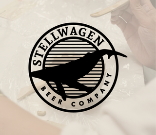 A circular black and white sticker or decal featuring the text 'STELLWAGEN BEER COMPANY' and a whale silhouette, shown over a softly blurred background with a hand holding a wooden spoon.