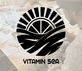 Black logo with 'Vitamin Sea' text on a blurred background of people doing pottery