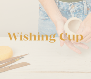 A person in blue jeans holds a small beige ceramic cup over a light wooden table. A yellow sponge and sculpting tools sit on the left, and the words 'Wishing Cup' are overlaid across the center.